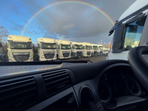 Looking out of a truck windscreen, a row of white HGV trucks with a rainbow over it.